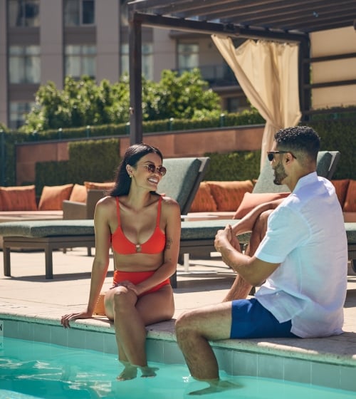 A man and a woman sitting on the edge of the rooftop pool at the Camby