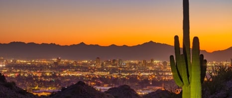Skyline of Midtown Phoenix at sunset with an orange skyline and mountains in the background and a mountainside and a partially lit Saguaro Cactus in the foreground.