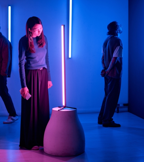 Individuals observing light sculptures in modern art gallery with neon blue and pink lighting creating an immersive, thoughtful environment