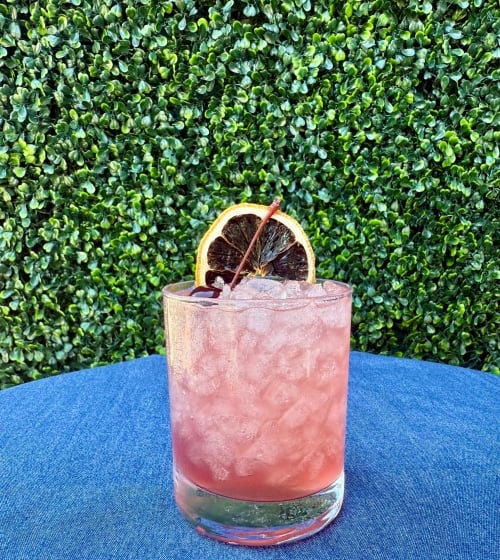 image of a pinkish purple cocktail with ice topped with a cherry garnish and dried lemon wheel
