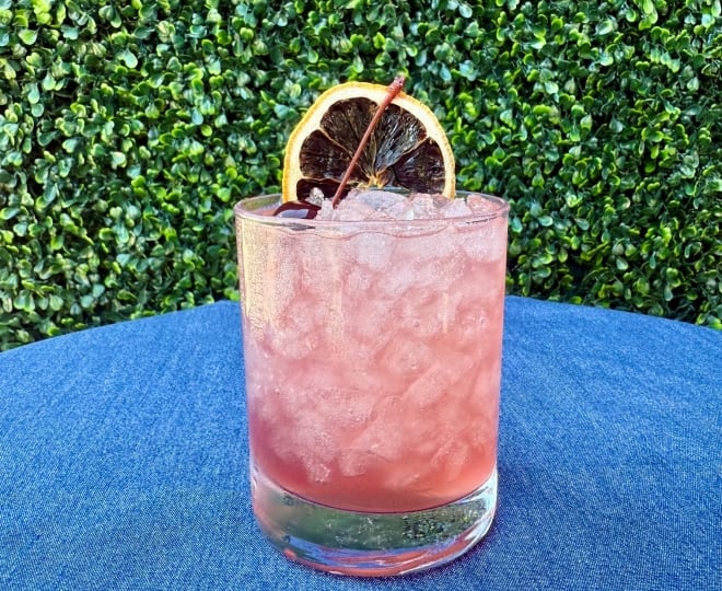 image of a pinkish purple cocktail with ice topped with a cherry garnish and dried lemon wheel